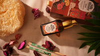 A bottle of Silk Route face serum with a gold pump and a decorative product box, styled on a beige fabric surface with dried pink flowers, a yellow sponge, chopsticks, and green leaves.