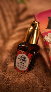 A bottle of Silk Route Face hydrating serum with a gold pump and amber glass bottle, resting on a textured brown surface with a pink box partially visible in the background.