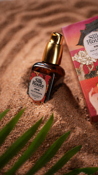 A bottle of Silk Route serum with a gold pump and amber glass, styled on a sandy surface next to a red product box and green leaves.