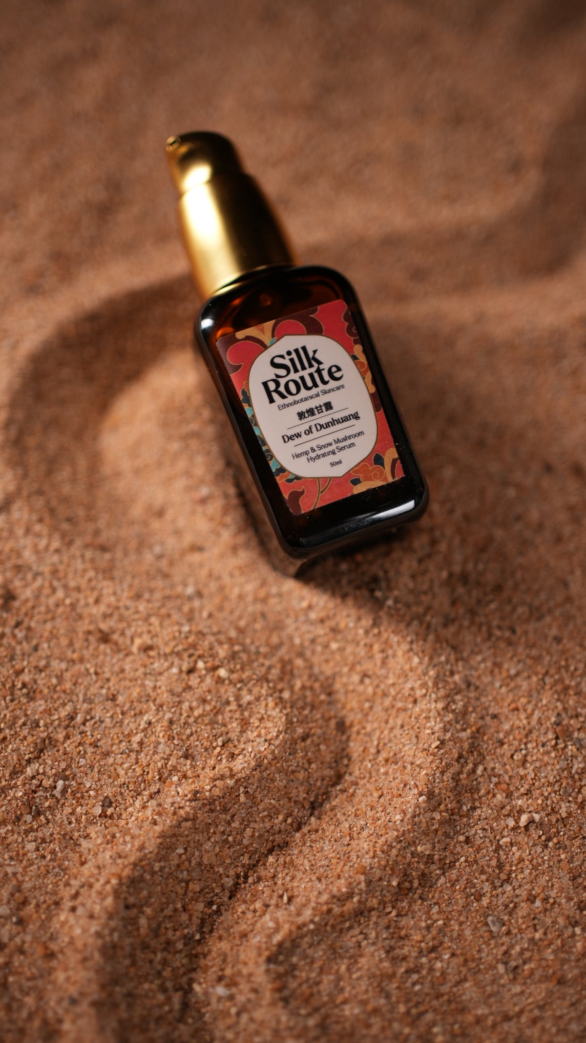 A bottle of Silk Route face serum with a gold cap and a red-and-white label, resting on a textured brown sand surface.