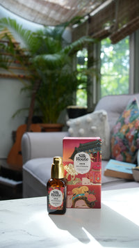 A bottle of hydraing serum and its qing dynatsy illustrated box are displayed on a white marble surface in a modern, sunlit room with a sofa and plants in the background.