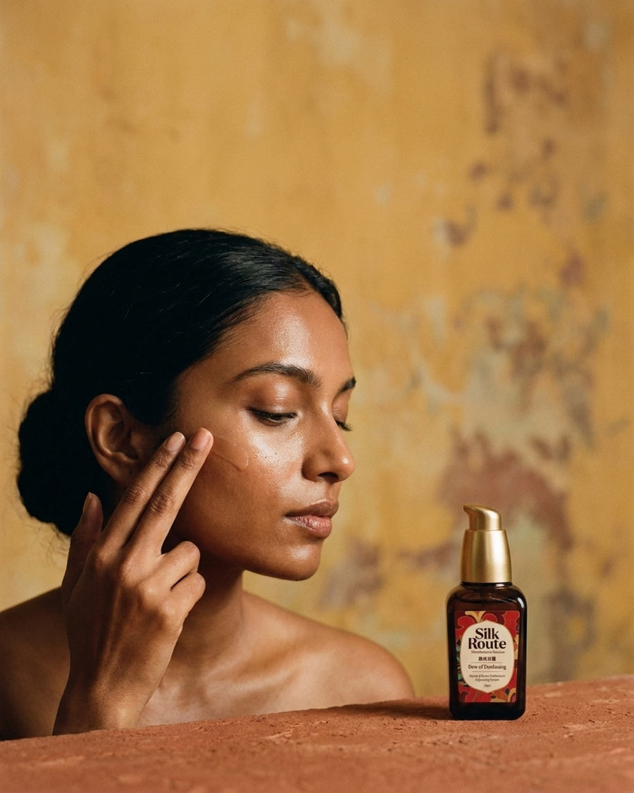A woman presses the Dew of Dunhuang serum into her cheek in front of an ochre fresco wall, the bottle in the foreground