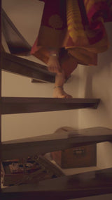 A woman in a red silk sari comes down the stairs at home, lights a brass lamp beside a Nataraja figure and the serum carton, and looks into a small round mirror.