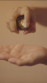 A woman presses a few drops of the Snow Mushroom Hydrating Serum from the bottle into her palm, then presses it into her cheeks, forehead and jaw in warm light.